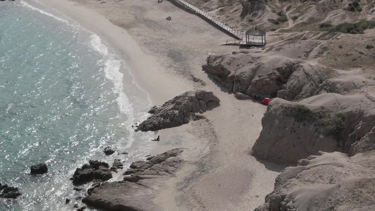 At 0:02, Playa El Arbolito is seen from high above with bright textured water, a narrow surf line, pale beach, rocky ledges, and small scattered beachgoers near the upper sand.