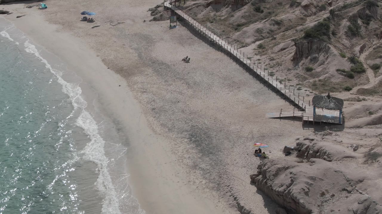 Aerial frame at Playa El Arbolito shows turquoise surf along pale sand, a fenced boardwalk crossing dunes, a thatched shade structure, umbrellas, and tiny beachgoers near the lower edge.