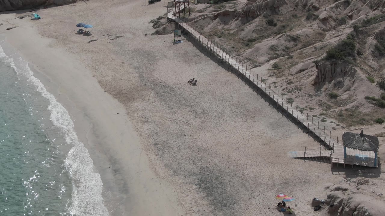 Aerial beach view with shoreline, boardwalk, dunes, umbrellas, and people on the sand.