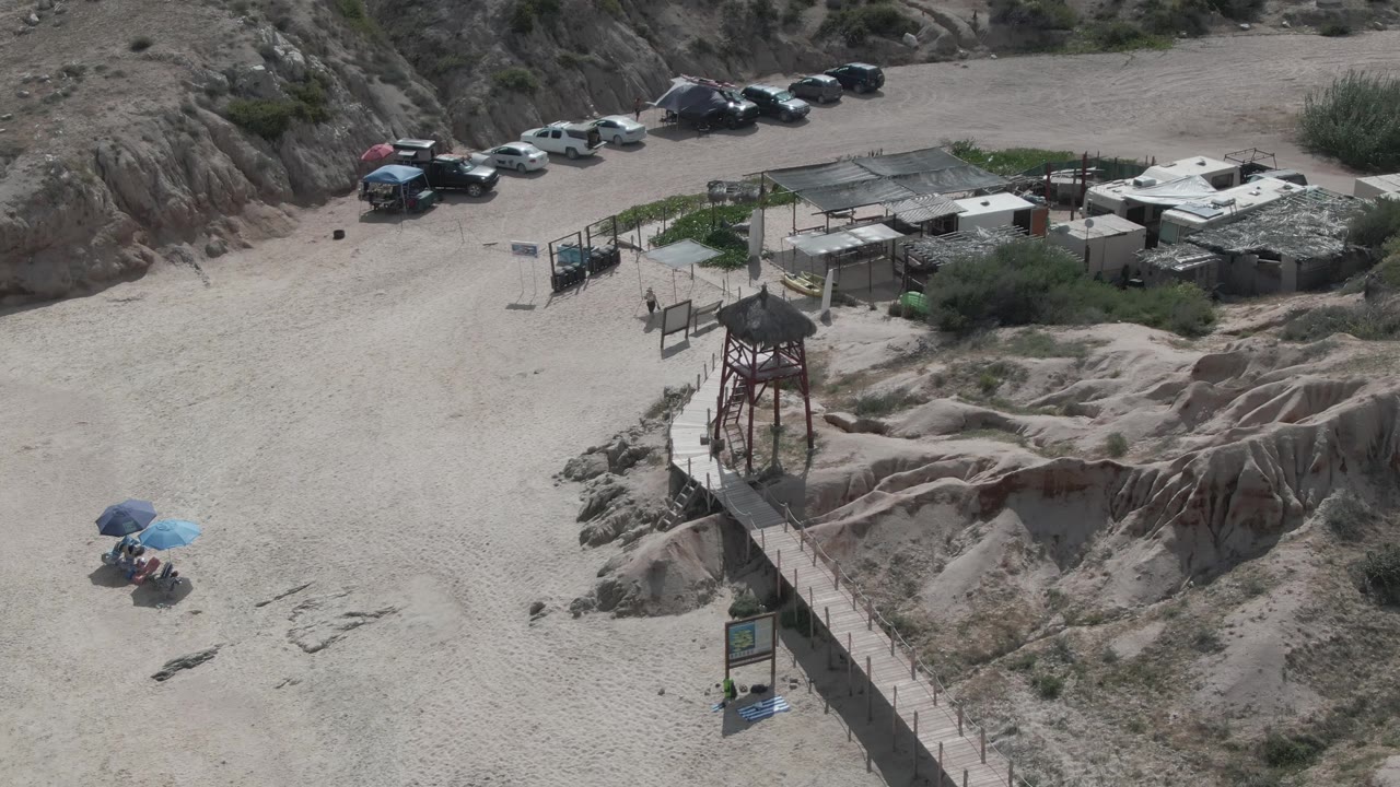 Aerial frame at Playa El Arbolito showing a sandy parking area with parked vehicles near rocky hills, a wooden walkway, a watchtower, small structures, umbrellas, and no clearly visible people.