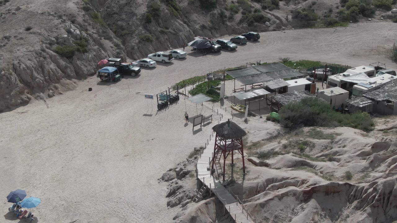Aerial frame at Playa El Arbolito with the watchtower lower in frame, small roofed structures beside the sandy lot, parked cars along the rocky slope, and no clearly visible people.