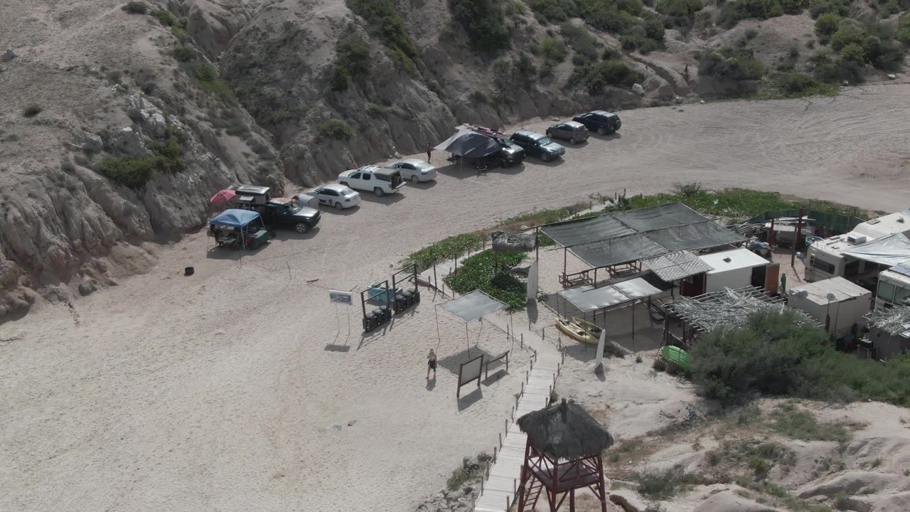 Aerial frame at Playa El Arbolito showing more of the sandy parking lot, a line of vehicles by the hillside, small buildings and fencing, with the watchtower partly near the lower edge and no clearly visible people.