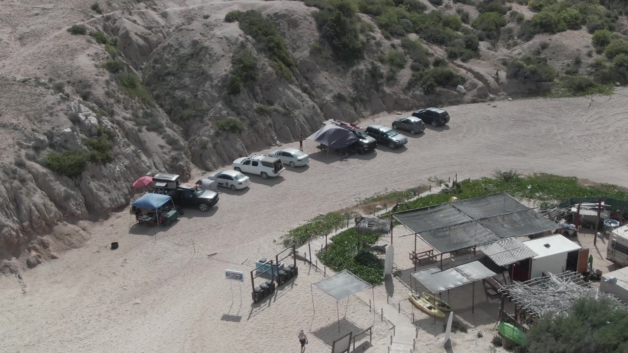 Aerial view of parked vehicles, shade umbrellas, small structure roofs, sandy ground, and rocky hillside.