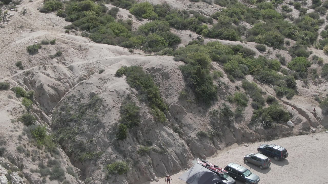 High-angle view at Playa El Arbolito Cabo Pulmo showing parked vehicles at the base of pale rocky hills with scattered green shrubs and no visible people.
