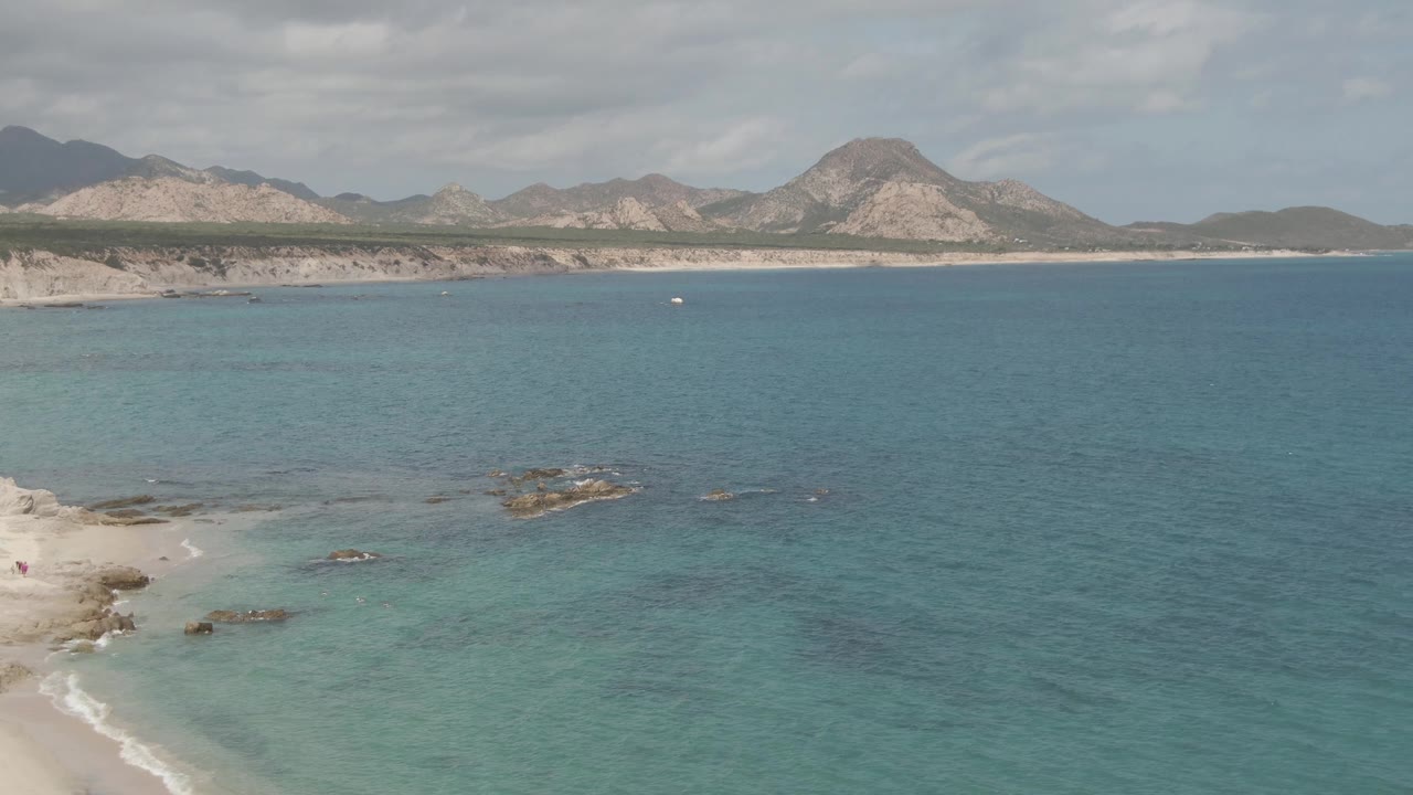 Aerial frame at Playa El Arbolito Cabo Pulmo BCS Mexico showing pale sand at the lower left, turquoise shallows, scattered rocks, a cliff line, and distant mountains under clouds.