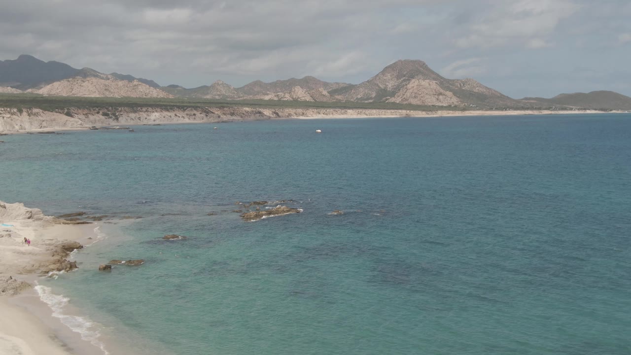 Wide aerial coastal view with beach corner, blue water, rocks, cliffs, and mountains.