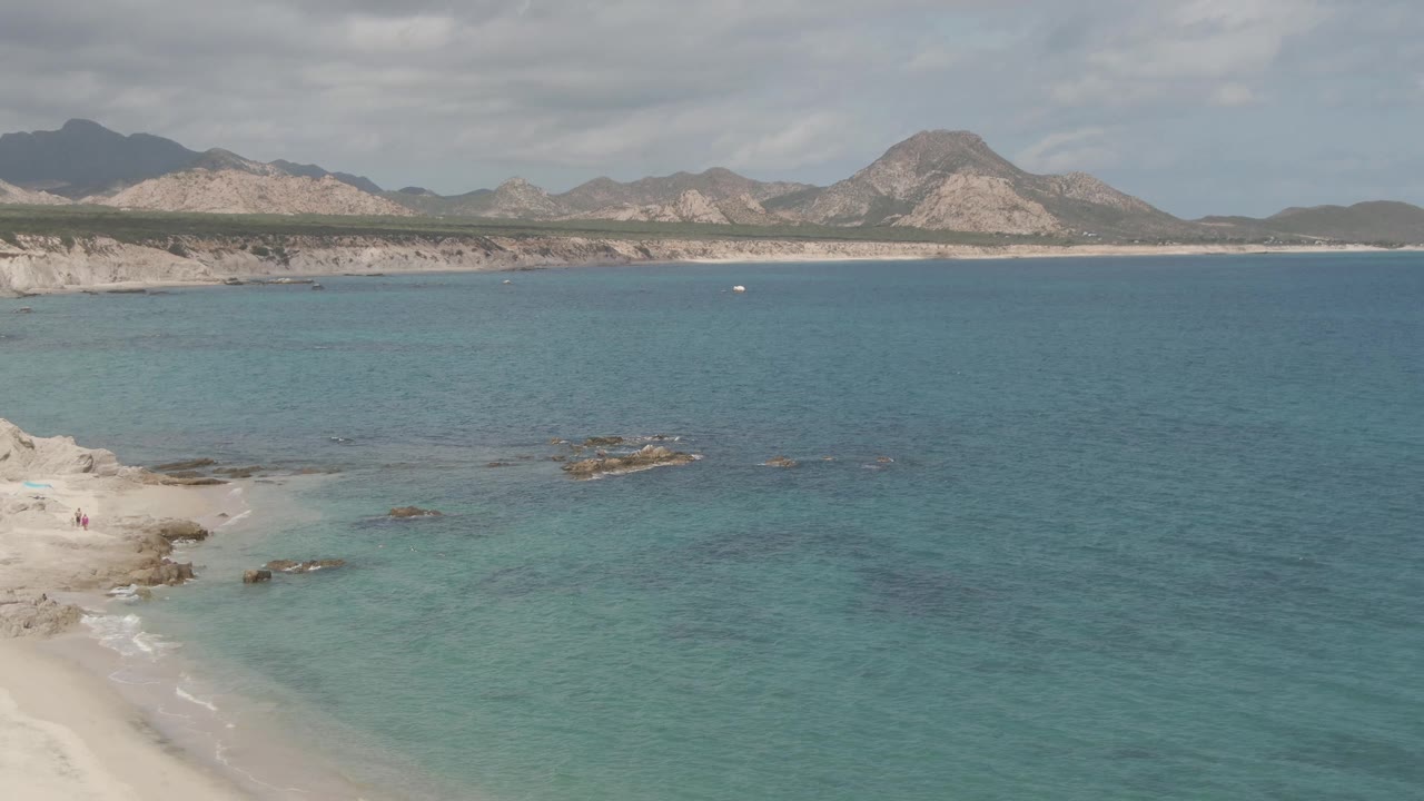 Frame from Playa El Arbolito Cabo Pulmo BCS Mexico showing more open turquoise water, a small sandy beach section at left, dark nearshore rocks, tan cliffs, and layered mountains.