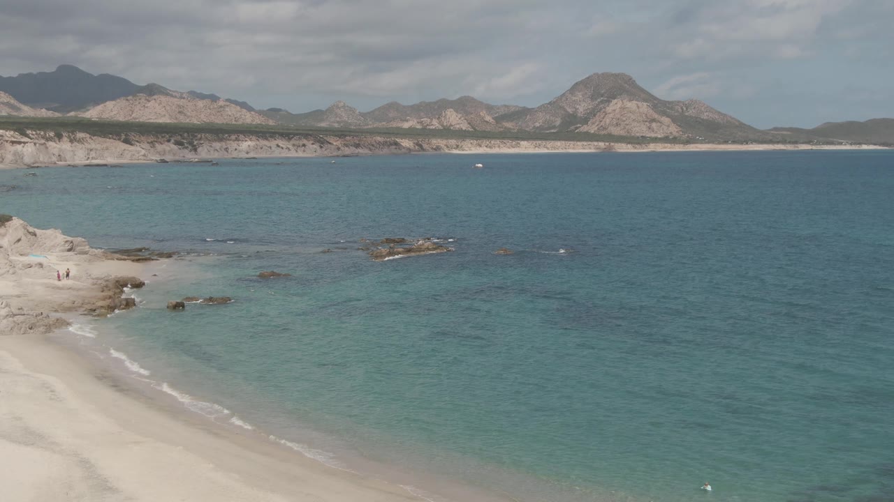 Elevated view over Playa El Arbolito with pale sand, turquoise water, shoreline rocks, distant mountains, and no visible people in the center of the frame.