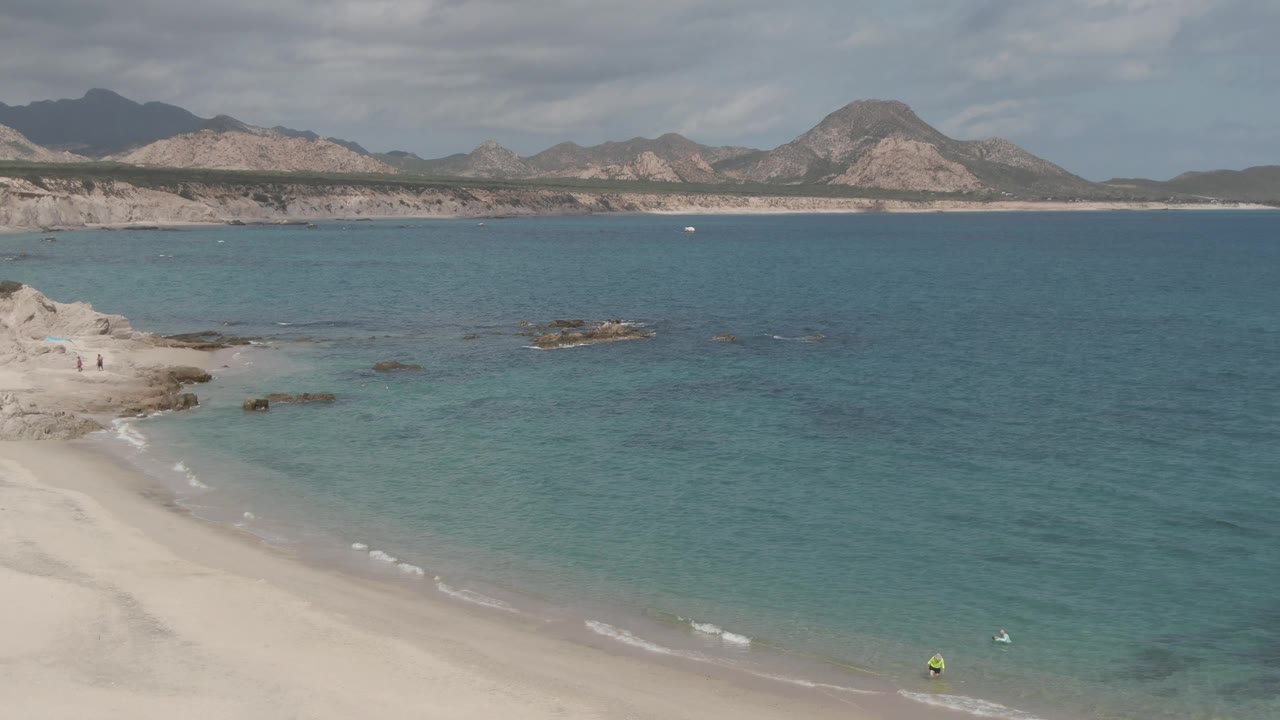 Wide Playa El Arbolito beach frame with clear water, a rocky outcrop near the center, small swimmers near the lower right, distant mountains, and a clouded sky.