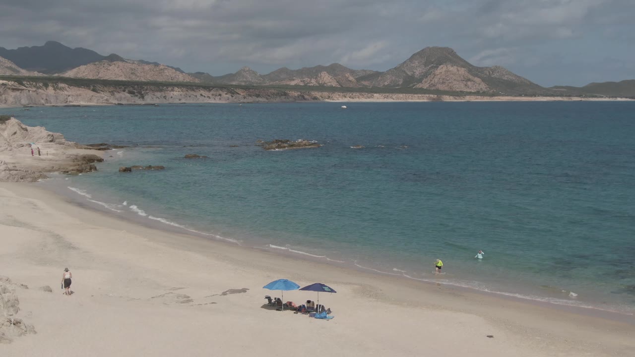 At Playa El Arbolito Cabo Pulmo, Mexico, an elevated view shows pale sand, a blue umbrella group, a person near the lower left beach, calm blue water, rocky outcrops, and mountains across the bay.