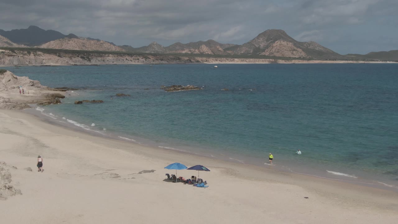 Sandy shoreline with people under blue umbrellas, a walker near rocks, and calm blue water.