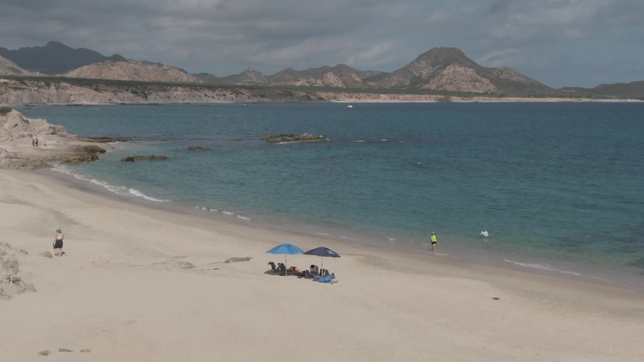 At 0:02, the elevated Playa El Arbolito Cabo Pulmo view keeps the umbrella group near the lower center, with a person in bright yellow near the waterline, small white waves, rocks offshore, and mountains beyond.