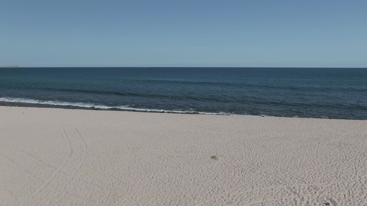 Opening frame of Buzzards Beach East Cape BCS Mexico with pale sand in the foreground, gentle surf at midframe, open ocean beyond, clear sky, and no visible people.