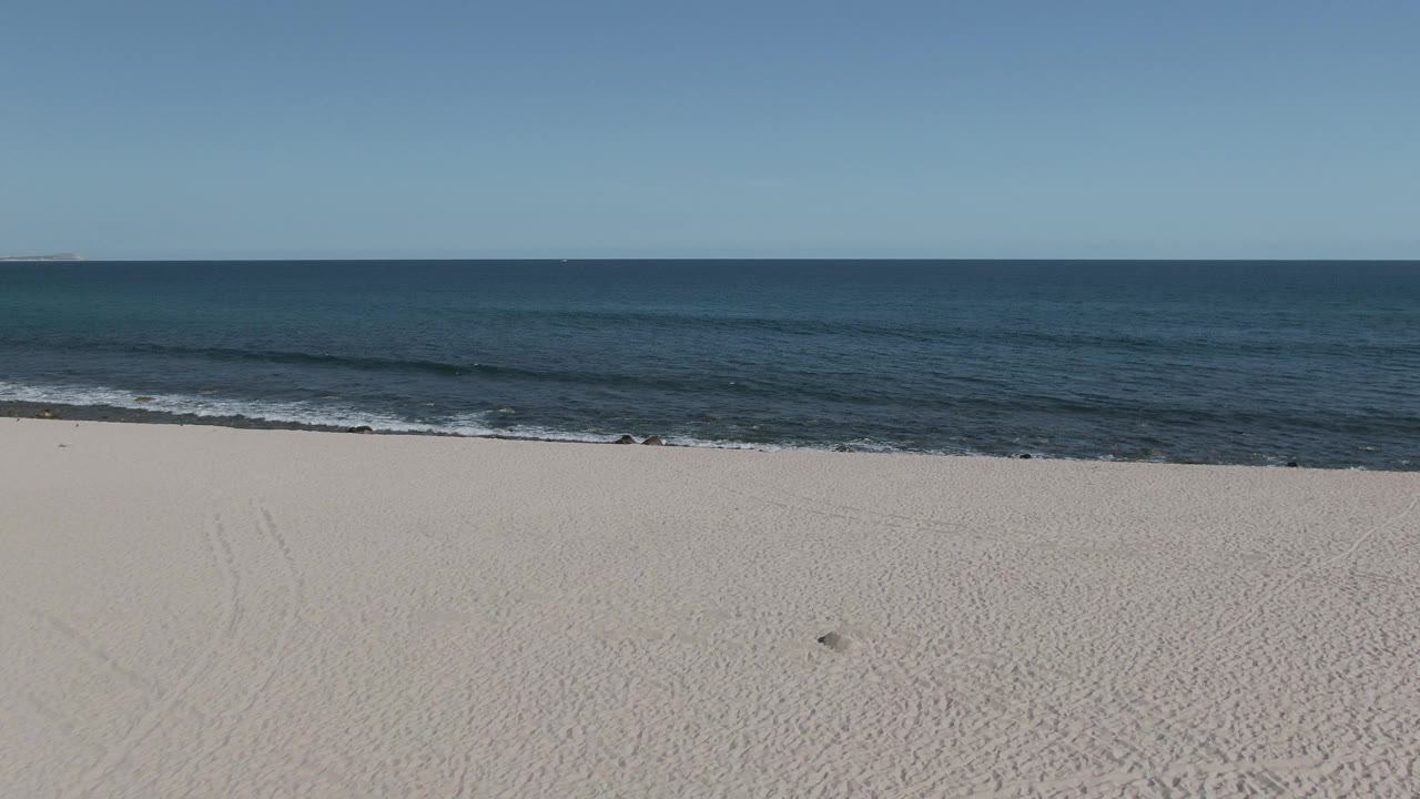 Buzzards Beach East Cape BCS Mexico shoreline at 0:01, showing smooth pale sand, a thin band of breaking waves, deep blue water, clear sky, and no visible people.