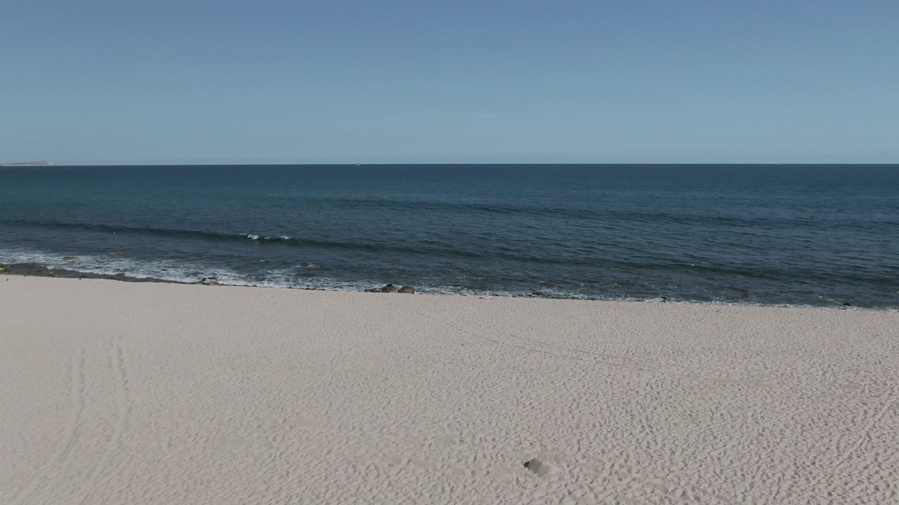 At 0:02 on Buzzards Beach East Cape BCS Mexico, the frame includes pale sand, a small dark rock near the shore, gentle waves, open blue water, clear sky, and no visible people.