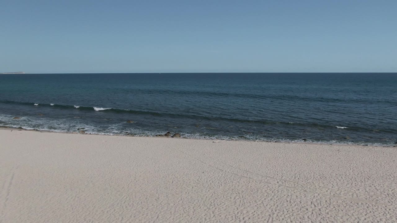 Low waves roll toward a wide sandy beach beneath blue sky.