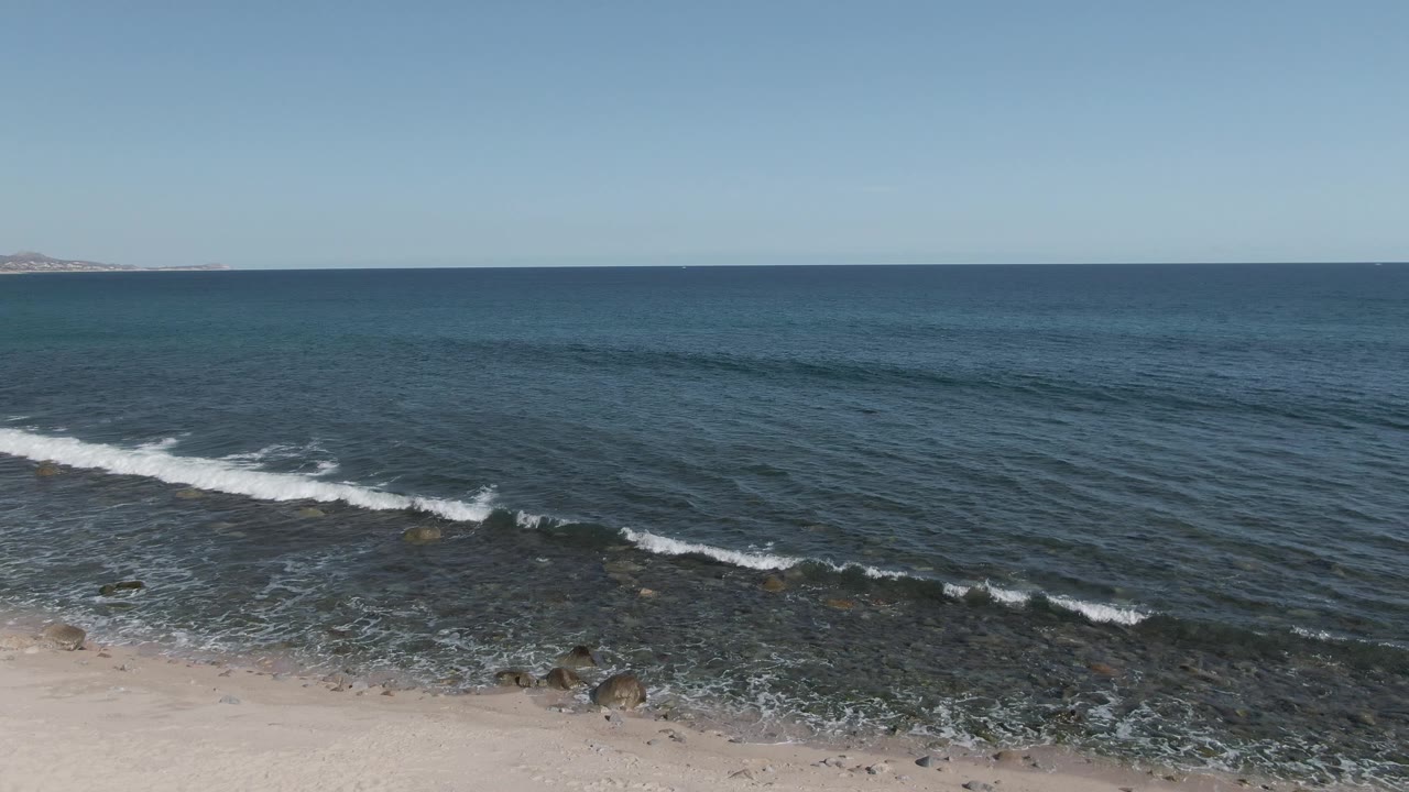 Wide aerial frame at Buzzards Beach East Cape BCS Mexico showing white foam pulling back from a sandy and rocky shoreline into clear blue water, with no visible people.