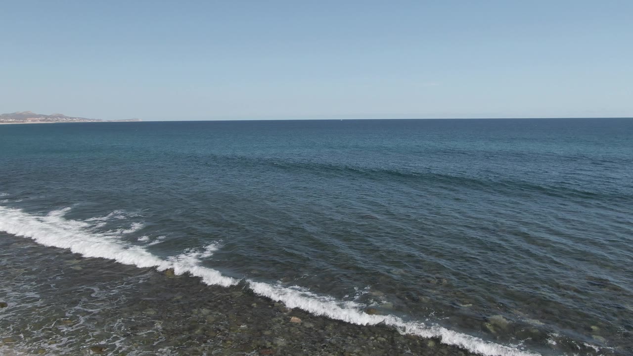 Aerial frame at Buzzards Beach East Cape BCS Mexico showing a low white wave line across blue water, rocky shallows below, distant land on the horizon, and no visible people.