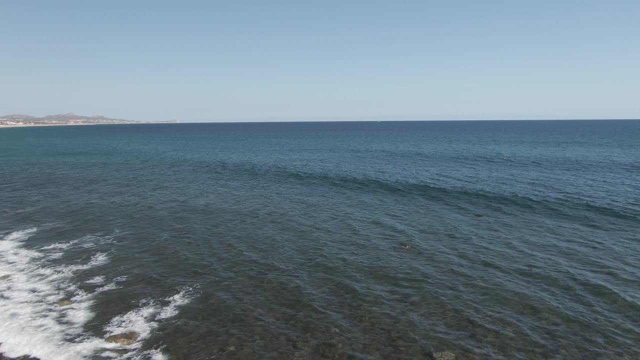 Aerial frame at Buzzards Beach East Cape BCS Mexico showing open blue ocean, white surf crossing shallow water near the foreground, a flat horizon, and no visible people.