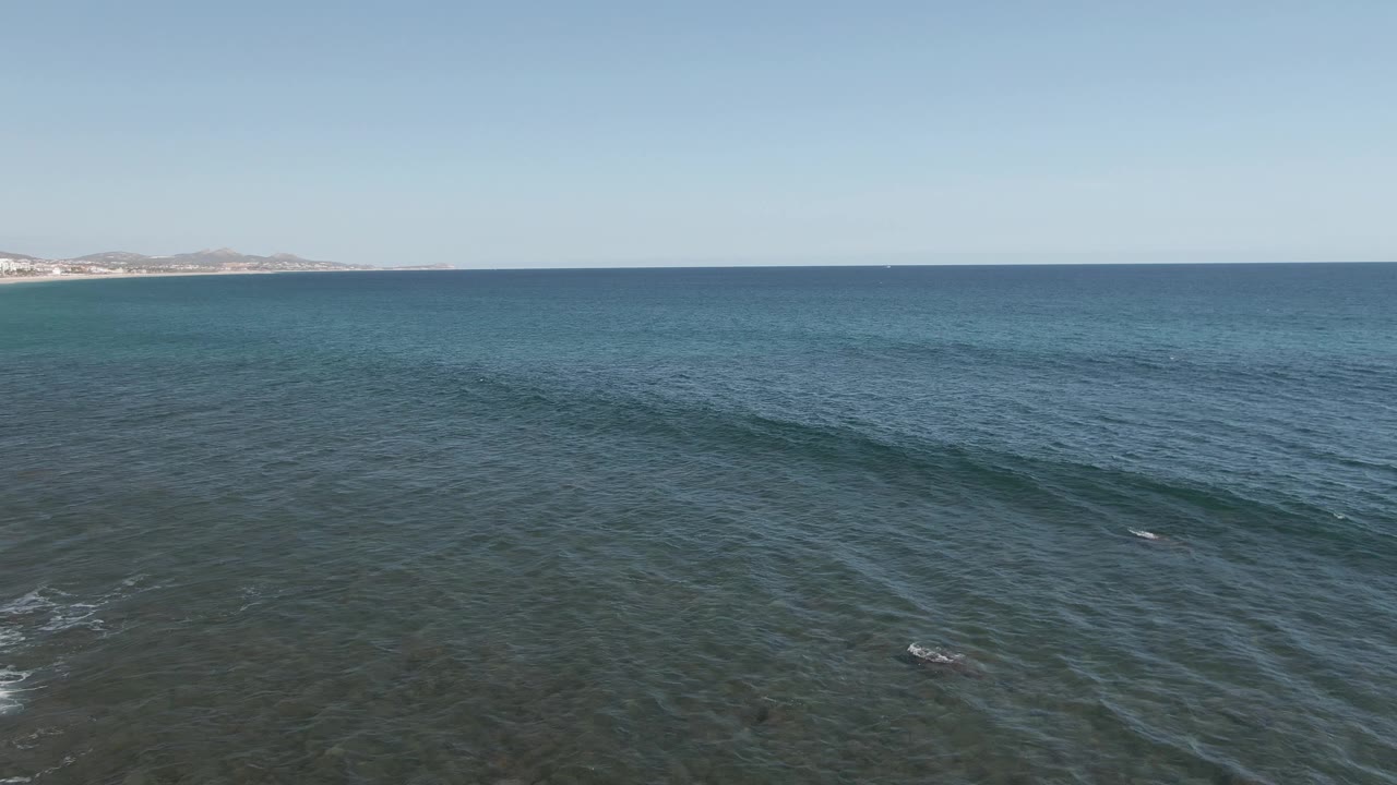 Aerial frame at Buzzards Beach East Cape BCS Mexico looking across turquoise and blue water toward a distant coastline and low hills, with no visible people.