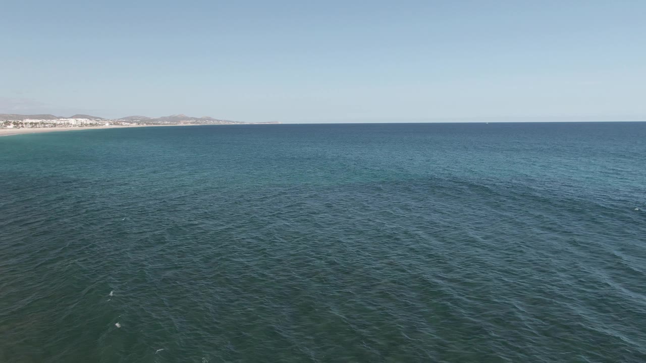 Opening aerial frame over blue-green ocean at Buzzards Beach East Cape BCS Mexico, with a sandy shoreline and low coastal buildings far left, distant hills, clear sky, and no visible people.