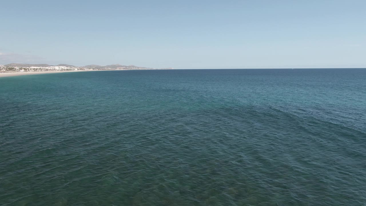 Aerial ocean view at Buzzards Beach East Cape BCS Mexico, with textured blue-green water in the foreground, the sandy coast and pale buildings farther left, and no visible people.