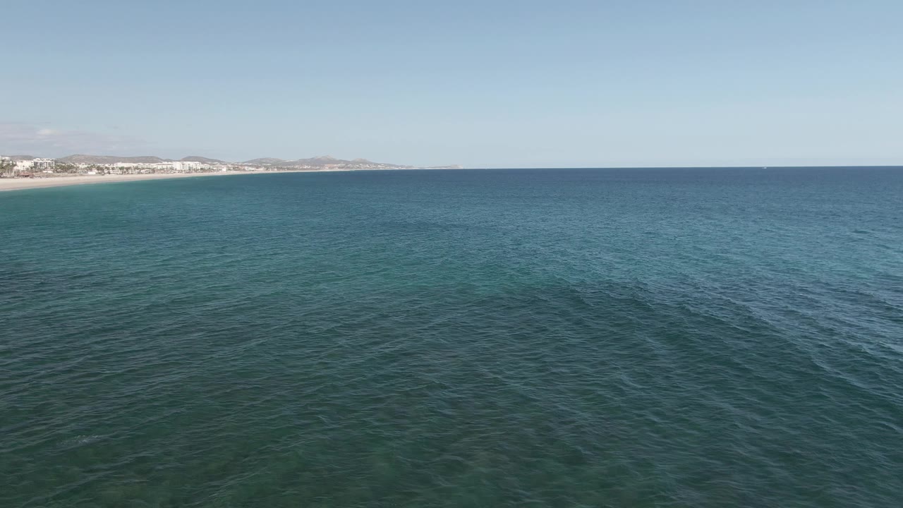 Wide drone-like frame over Buzzards Beach East Cape BCS Mexico, showing darker blue-green water bands, distant sandy shoreline, low buildings at left, dry hills, and no visible people.