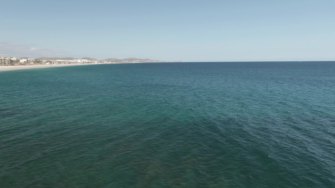 Mid-clip aerial frame at Buzzards Beach East Cape BCS Mexico, with the sandy beach and coastal buildings slightly more prominent on the left and calm blue-green water across the foreground, with no visible people.
