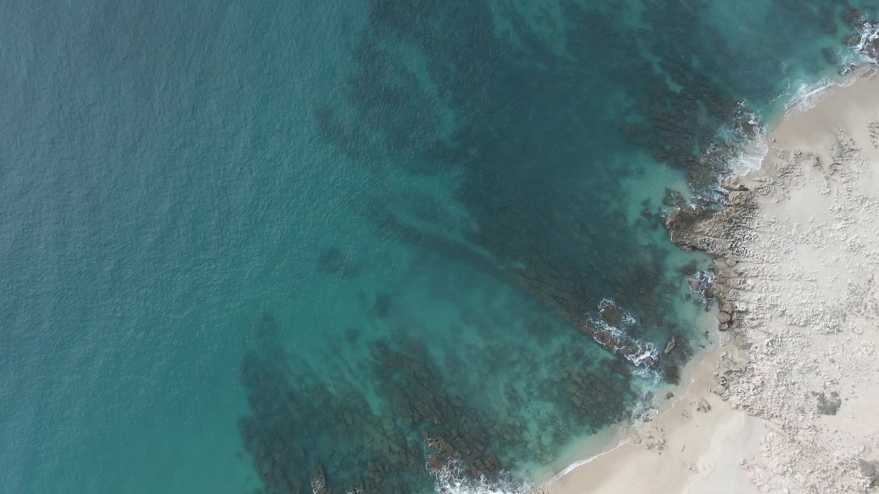 Aerial frame of clear teal water over dark rock patches beside a pale sandy edge at Buzzards Beach East Cape BCS Mexico, with no visible people.
