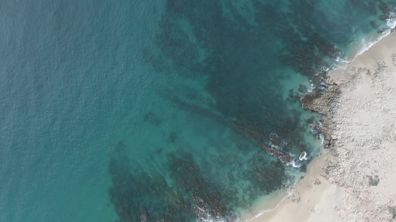Later overhead frame of teal ocean water spreading across dark submerged rocks and a sandy shoreline at Buzzards Beach East Cape BCS Mexico, with no visible people.