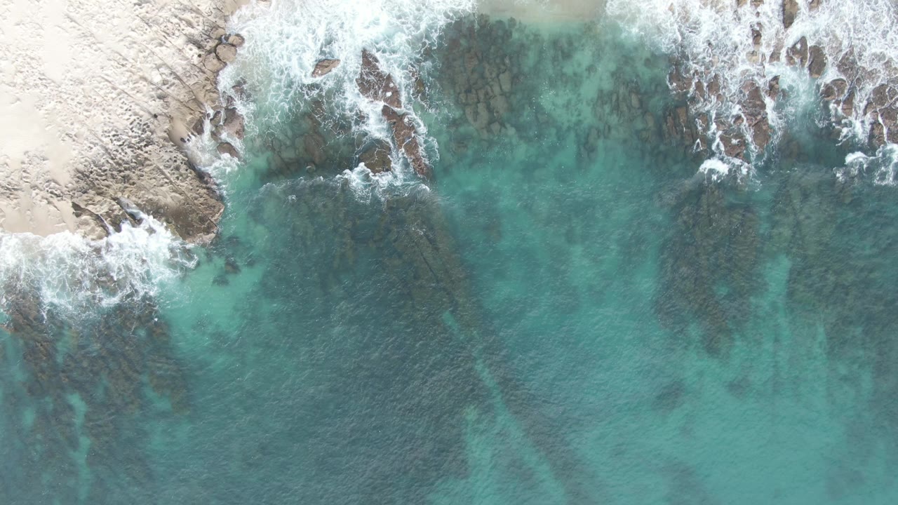 Overhead frame from Buzzards Beach East Cape BCS Mexico showing turquoise water, dark submerged rocks, white foam at the shoreline, and no visible people.