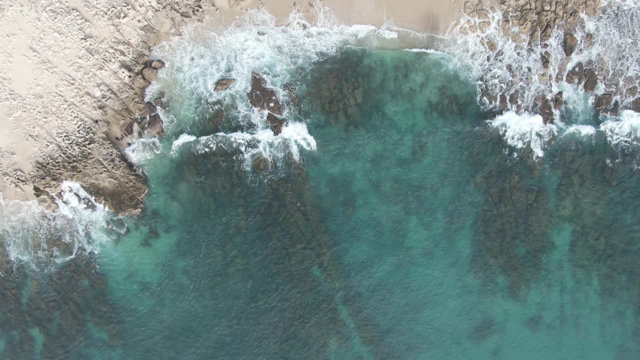Top-down coastal frame from Buzzards Beach East Cape BCS Mexico with the surf line pushed against a pale rocky sand edge and no visible people.