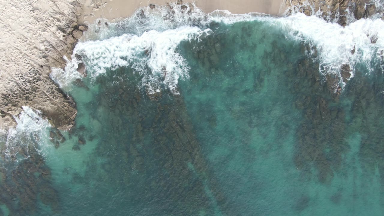 Overhead frame from Buzzards Beach East Cape BCS Mexico showing white surf at the pale rocky shore, turquoise water over dark submerged rocks, and no visible people.