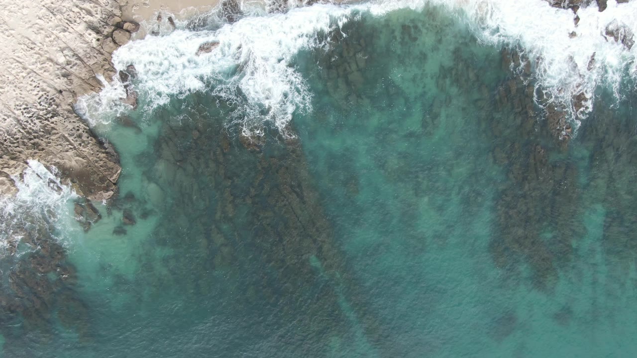 At 0:01, white foam pushes along the upper shoreline while green-blue water reveals darker rock streaks below the surface at Buzzards Beach East Cape BCS Mexico, with no visible people.