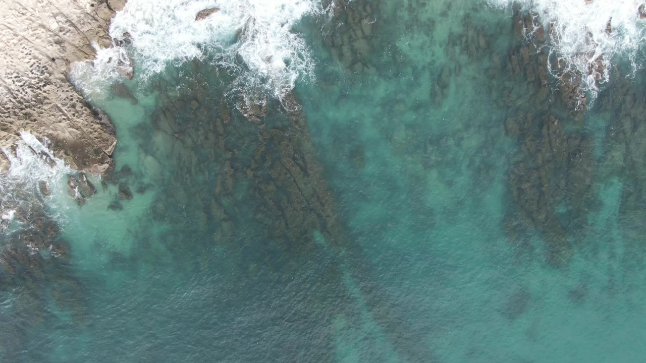 At 0:02, foamy surf breaks across the top edge of the rocky shore while transparent turquoise water exposes long dark rock lines beneath, with no visible people.