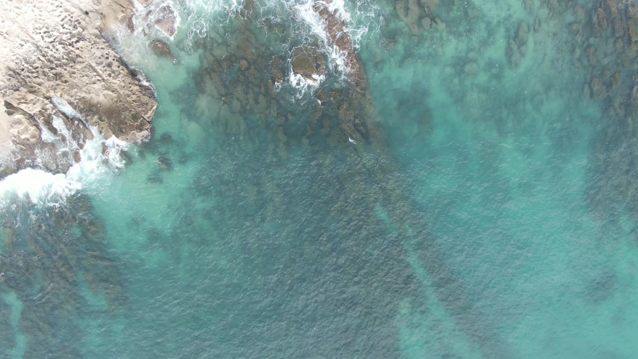 At Buzzards Beach East Cape BCS, Mexico, a top-down aerial frame shows turquoise water, submerged rocks, pale shoreline at upper left, white surf, and no visible people.