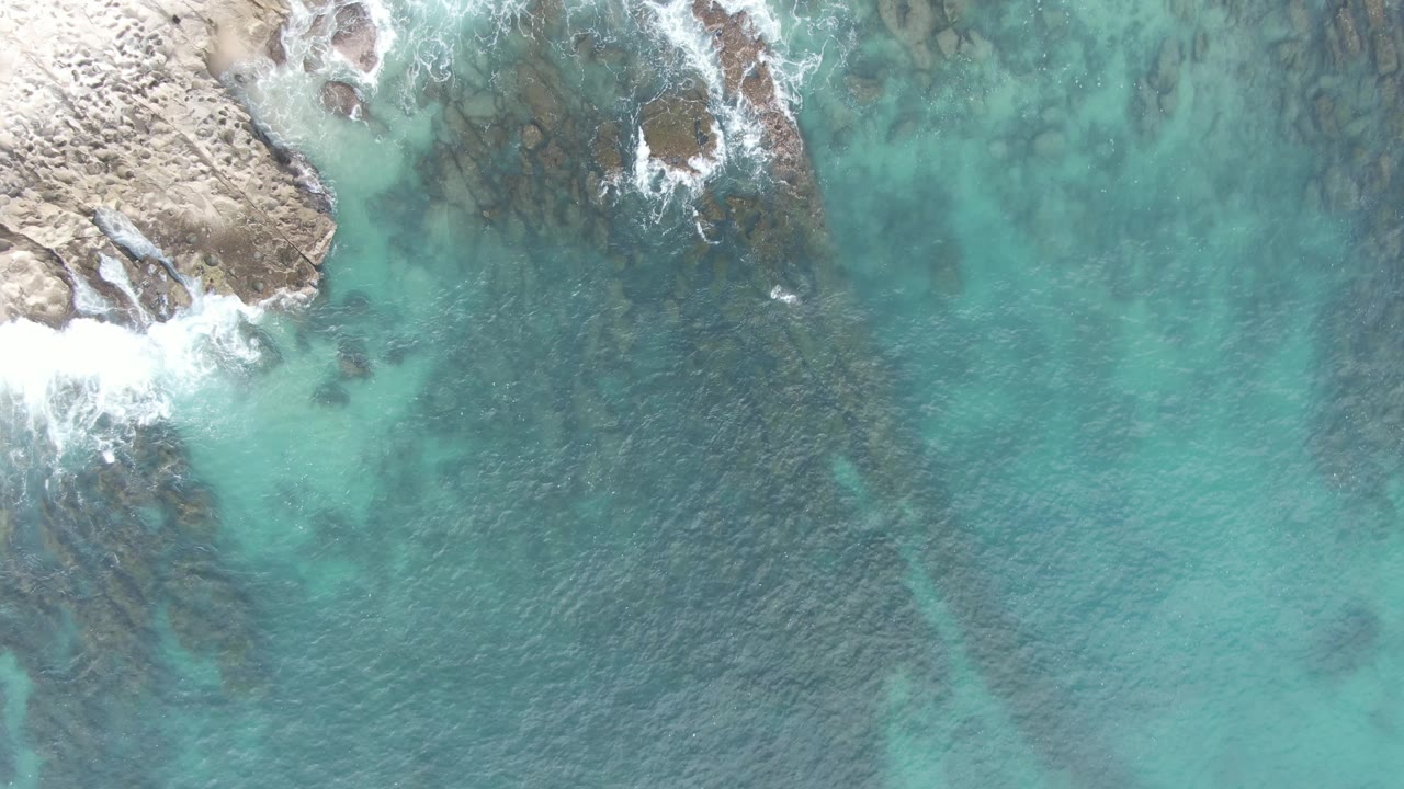 At 0:01, the Buzzards Beach aerial view holds the rocky shore near the upper left while clear blue-green water reveals dark submerged rock bands and no visible people.