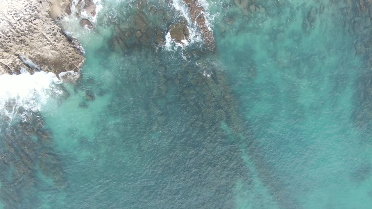 At Buzzards Beach East Cape BCS, Mexico, white surf spreads along the rocky upper-left shore while transparent turquoise water covers darker rocks below, with no visible people.