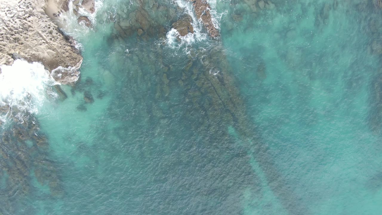 Drone view of rocky coast, foam, and shallow turquoise water with underwater rocks.