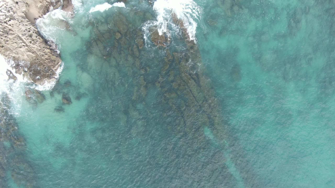Overhead view of turquoise water, submerged rocks, and foam beside a rocky shore.