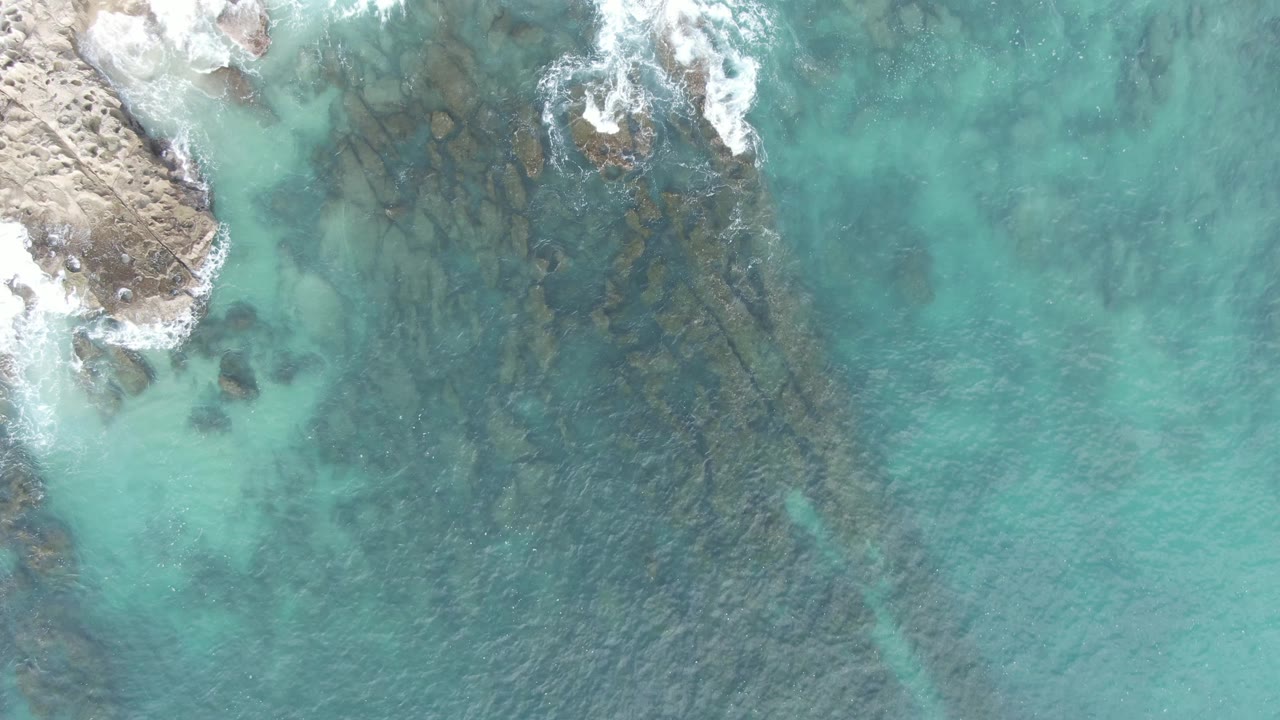 Overhead aerial frame at Buzzards Beach East Cape BCS Mexico with breaking white foam near the top edge, rocky coast on the left, darker submerged ledges through the center, and no visible people.