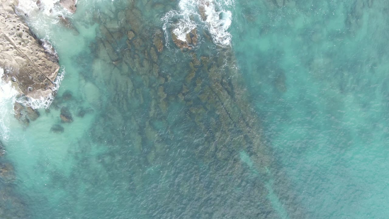 Overhead aerial frame at Buzzards Beach East Cape BCS Mexico where the rocky point stays at the left edge, foam gathers along the upper rocks, clear water shows dark submerged shapes, and no visible people.
