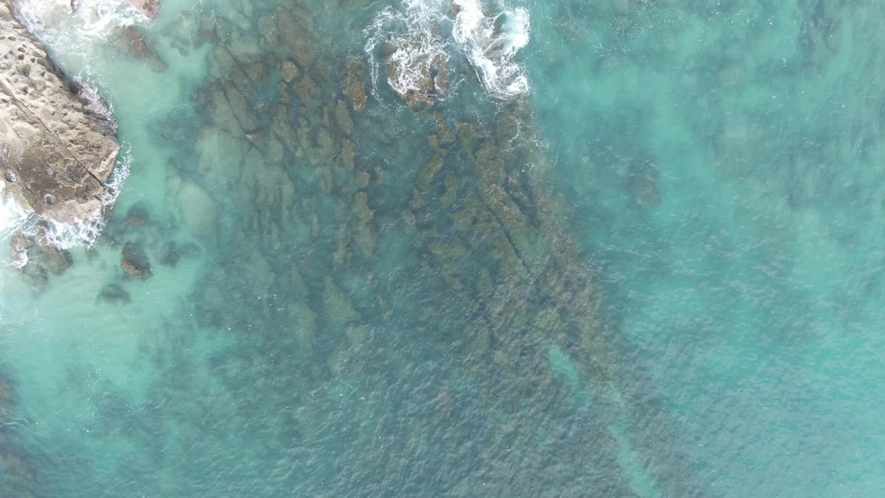 Overhead aerial frame at Buzzards Beach East Cape BCS Mexico showing a slightly lower moment in the shoreline pass, with foamy water at the upper left, submerged rocks spread through the center, and no visible people.