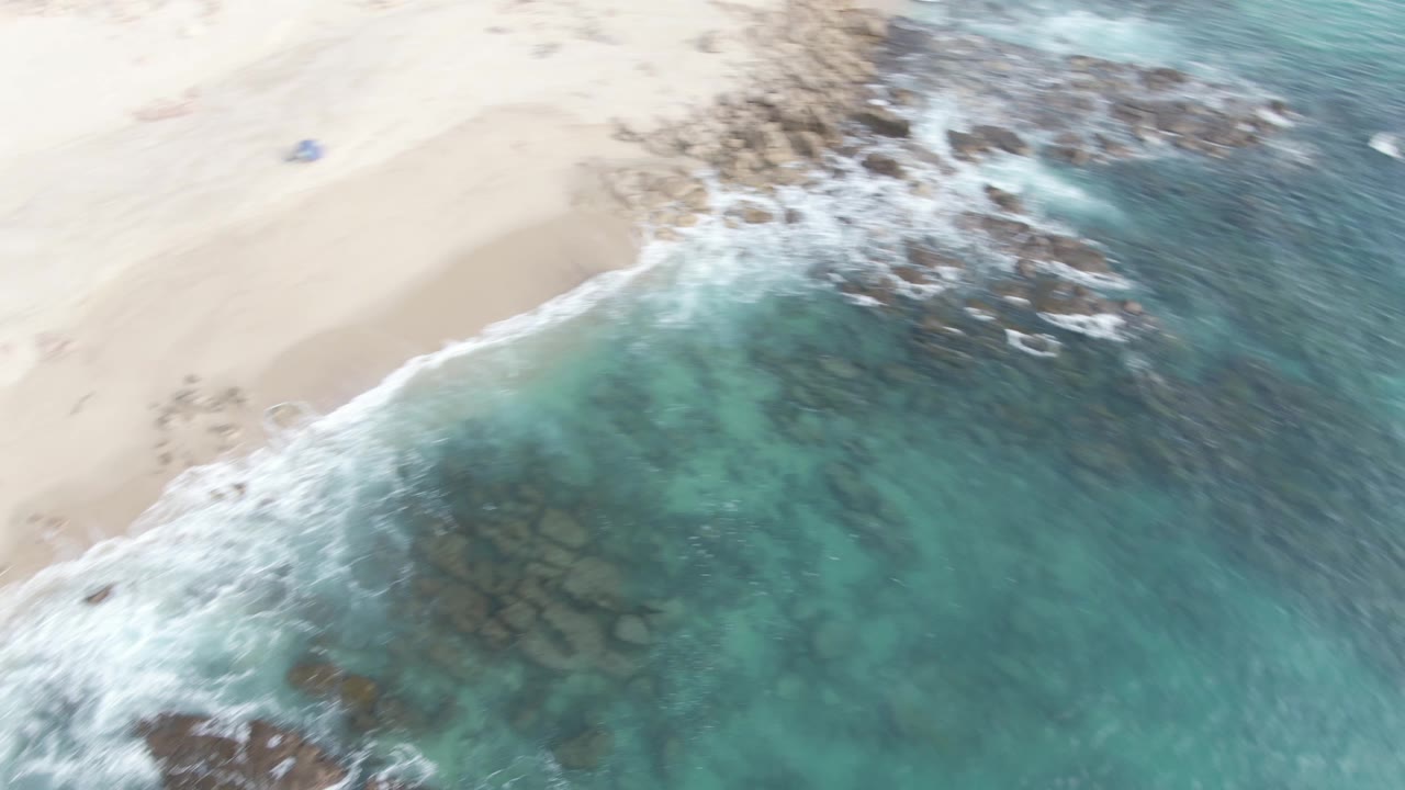Aerial frame over Buzzards Beach East Cape BCS Mexico showing pale sand, dark rocks, turquoise surf, and no visible people.