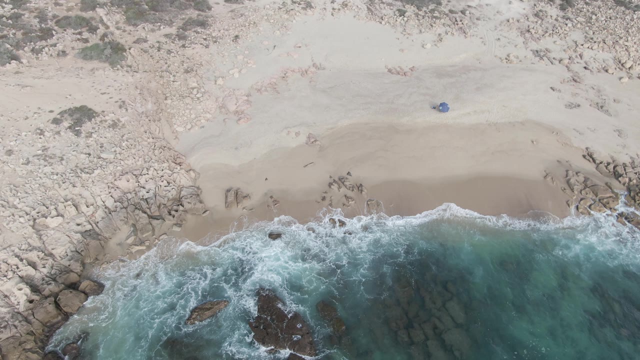 Top-down frame at Buzzards Beach East Cape BCS Mexico with a wide strip of sand, scattered rocks, breaking white foam, and no visible people.