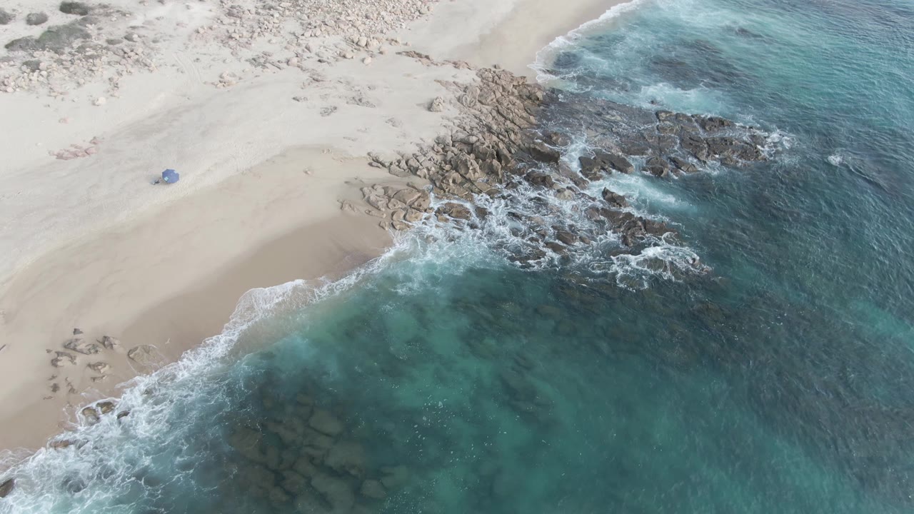 Aerial view along Buzzards Beach East Cape BCS Mexico showing clear blue-green water wrapping around a rocky point, pale sand above, and no visible people.