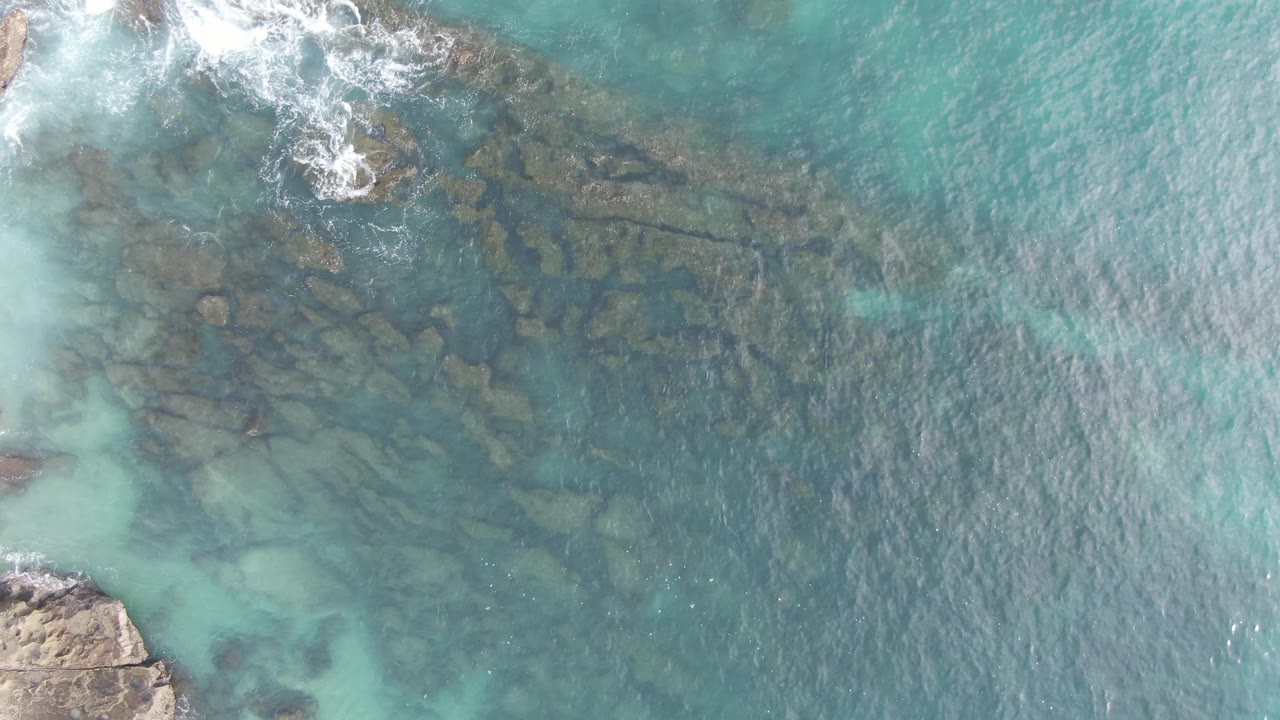 Overhead view at Buzzards Beach East Cape BCS Mexico showing turquoise water, submerged rocky seabed, white foam near the upper left, and no visible people.
