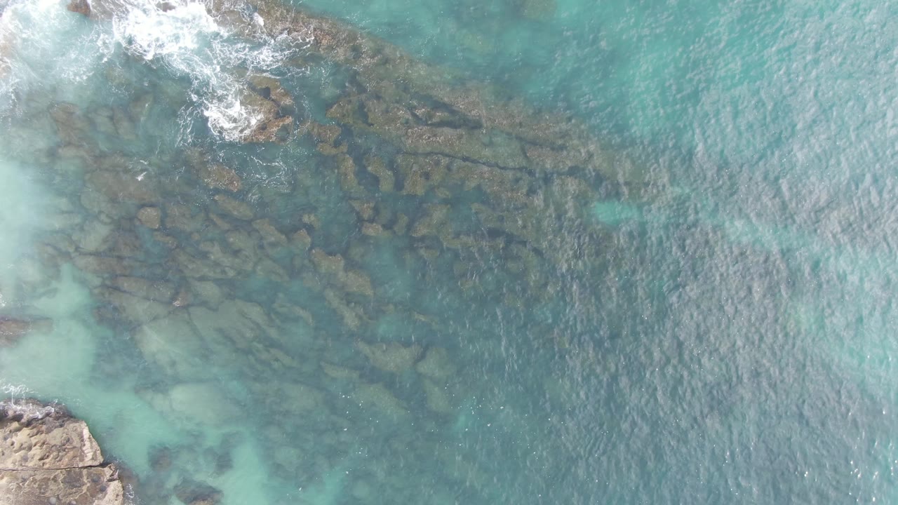 Aerial frame from Buzzards Beach East Cape BCS Mexico with dark underwater rock formations below teal water, bright foam along the top edge, and no visible people.