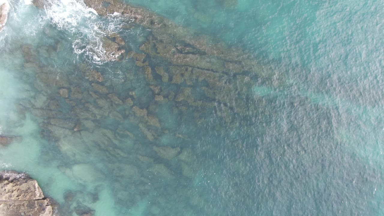 Top-down Buzzards Beach East Cape BCS Mexico view where a band of white surf crosses the upper frame above submerged rocks and open teal water, with no visible people.