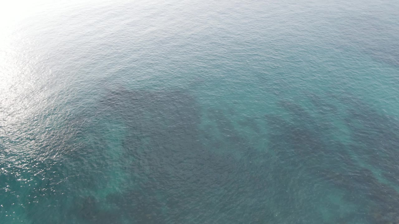 Sunlit aerial frame at Buzzards Beach East Cape BCS Mexico with bright reflection on the water, turquoise shallows, darker underwater reef areas, and no visible people.